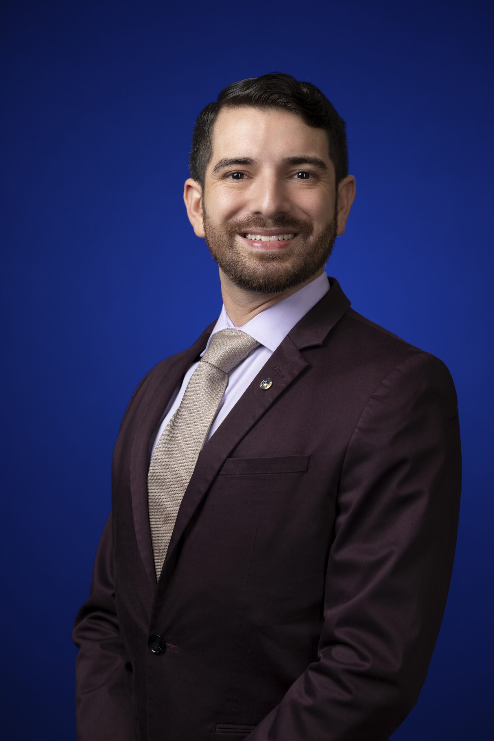 Portrait of 2025-2026 District Governor John Cano. He is in business professional dress with a white shirt, gold tie, and eggplant colored blazer against a blue background.